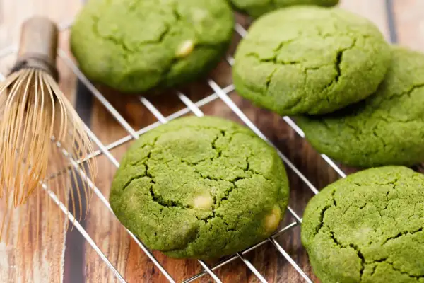 Zielone, japońskie ciasteczka matcha z kawałkami białej czekolady na metalowej kratce, obok bambusowej miotełki do herbaty.