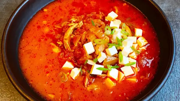 Pikantna zupa kimchi z kurczakiem i kostkami tofu, udekorowana szczypiorkiem.