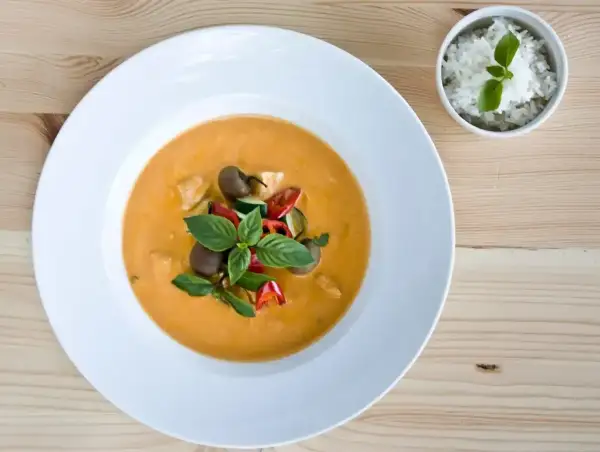 Aksamitny **thai red curry** z kurczakiem, cukinią i pomidorkami, udekorowany bazylią. Obok miseczka ryżu.