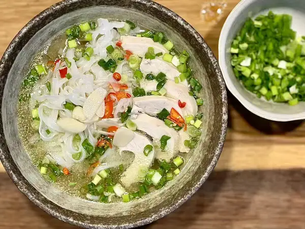 Aromatyczna zupa pho ga z kawałkami kurczaka, makaronem ryżowym, szczypiorkiem i papryczką chili. Obok miseczka z posiekanym szczypiorkiem.