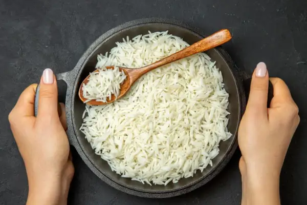 Dwie dłonie trzymają okrągłą patelnię z parującym ryżem basmati. Drewniana łyżka zanurzona w ryżu.