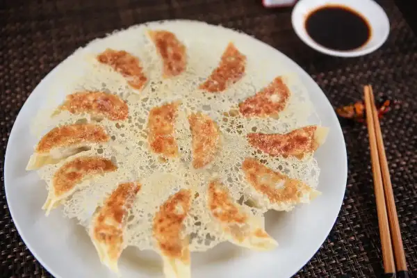 Chrupiące pierożki gyoza ułożone na talerzu, z chrupiącą panierką z ciasta. Obok miseczka sosu sojowego i pałeczki.