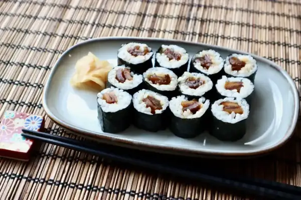 Tykwa sushi, czyli rolki z ryżem i ciemnym nadzieniem, ułożone na talerzu obok imbiru i pałeczek.