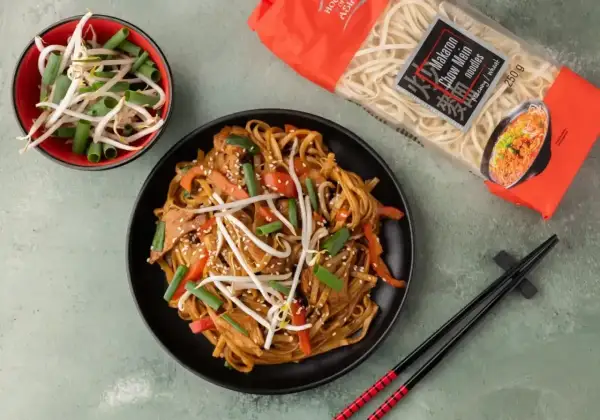 Chow mein z kurczakiem, warzywami i kiełkami fasoli mung, obok opakowania makaronu i miseczki z kiełkami. Idealne danie z wykorzystaniem przypraw chińskich.