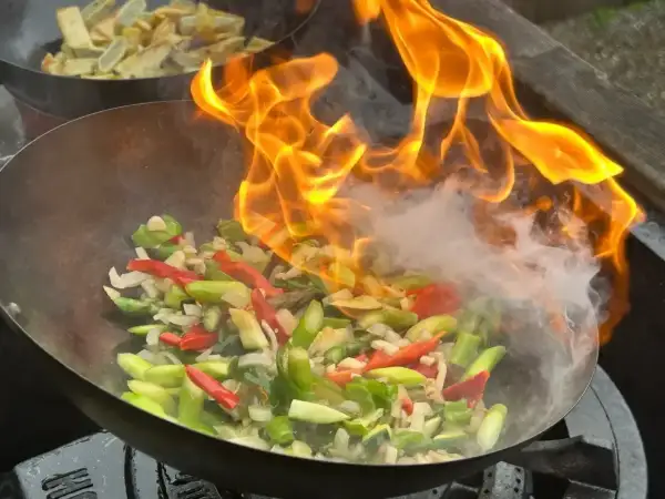 Płomienie otaczają warzywa w woku, tworząc apetyczny stir fry. W tle widać drugie danie.