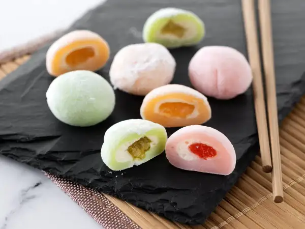 Kolorowe ciasteczka mochi z owocowym nadzieniem, ułożone na czarnej desce, obok pałeczek.
