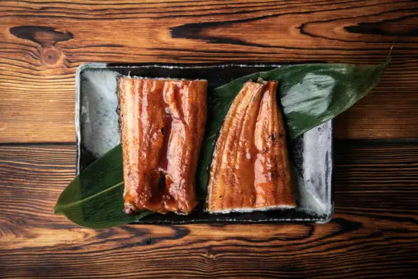 Dwa kawałki lśniącego, glazurowanego unagi na liściu, podane na kamiennej tacy na drewnianym tle.